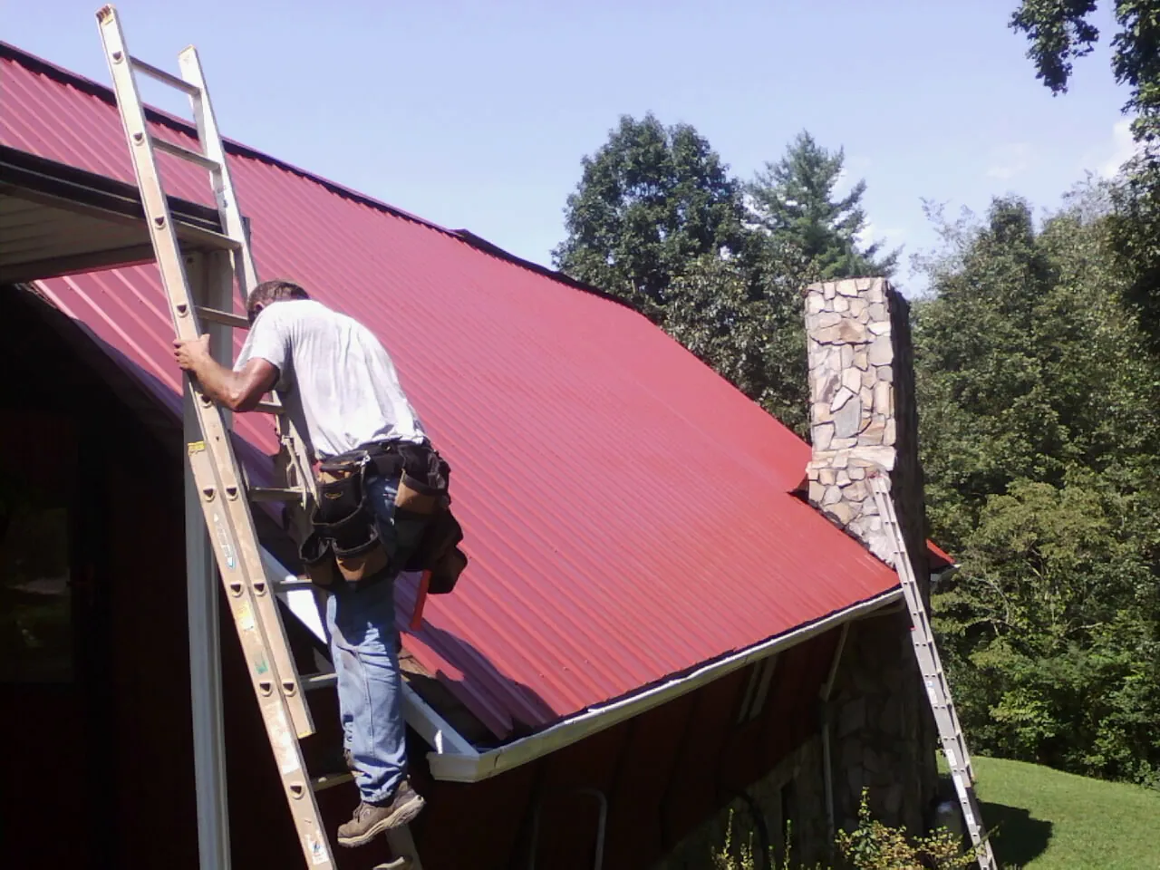 Roofer on a ladder beside a red metal roof and stone chimney — Soffit & Fascia Repair by Roofing Brothers in Charlestown